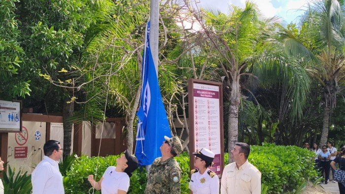 A group of people in white uniforms and a person in camouflage stand looking up at a blue flag being raised on a pole beside a sign listing beach rules, with palm trees and greenery in the background.