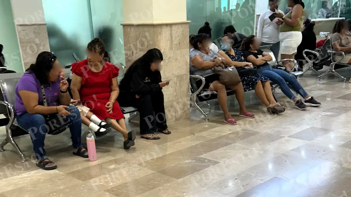 Several people sit on connected metal chairs in a tiled hall, some looking at phones, with others standing in the background.