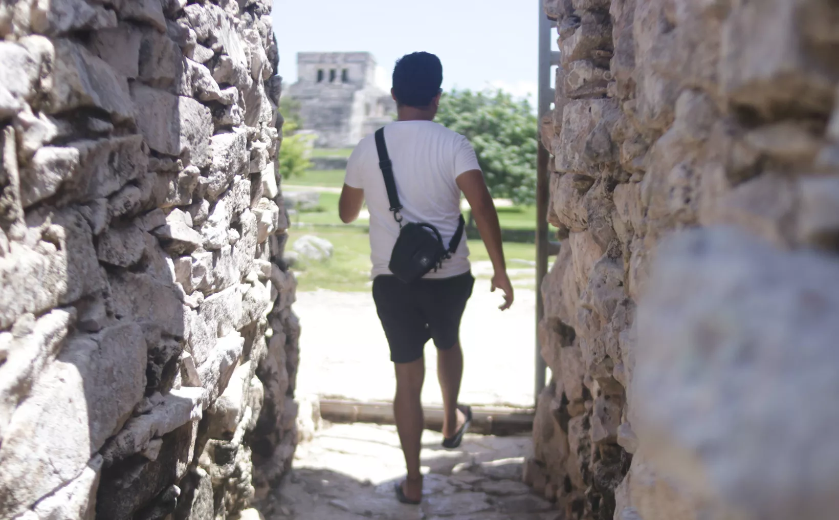 A person in a white t-shirt and black shorts with a black crossbody bag walks away from the camera through a narrow passage between rough stone walls toward a stone pyramid structure in the distance.