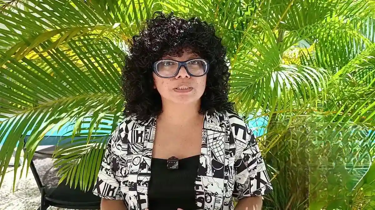 A person with curly black hair and clear-framed glasses wearing a black and white comic print shirt stands in front of palm fronds.
