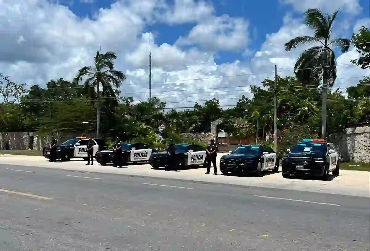 Five black and white police vehicles marked POLICIA are parked along the shoulder of a paved road, with several officers standing beside them, palm trees and a stone wall behind, under a blue sky with clouds.