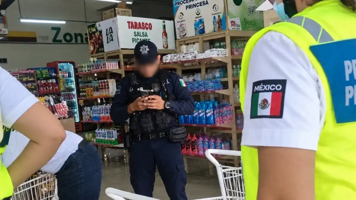 A police officer in dark uniform and cap stands in a grocery store aisle looking at a phone, with a person in a yellow vest and Mexico patch in the foreground.