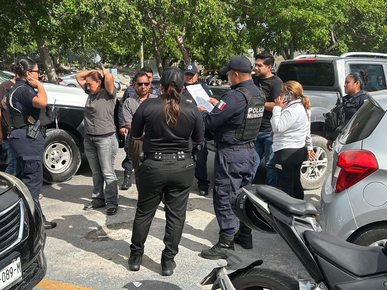 A group of police officers in dark uniforms and civilians stand on a street near parked vehicles, with one officer holding a document and a woman with her hands on her head.