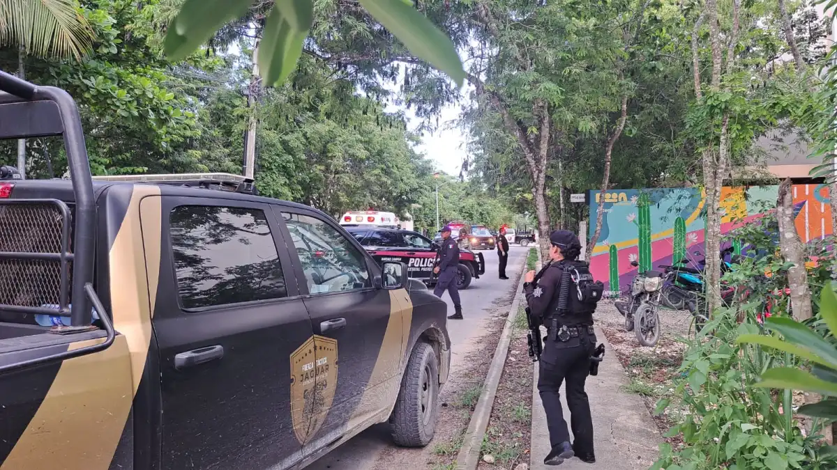 A black and tan police pickup truck is parked on a paved road beside a sidewalk where an officer in dark uniform stands, with other police vehicles and officers further down the street near a colorful mural.
