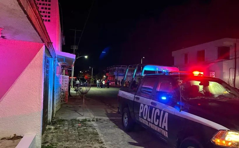 A police pickup truck with red and blue emergency lights illuminated is parked on a street at night beside a pink lit building, with people gathered in the background.