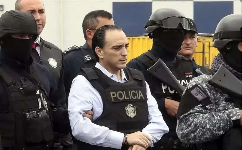 A group of armed police officers surround a man in handcuffs, who is wearing a police vest. The scene appears to be a police operation or arrest.