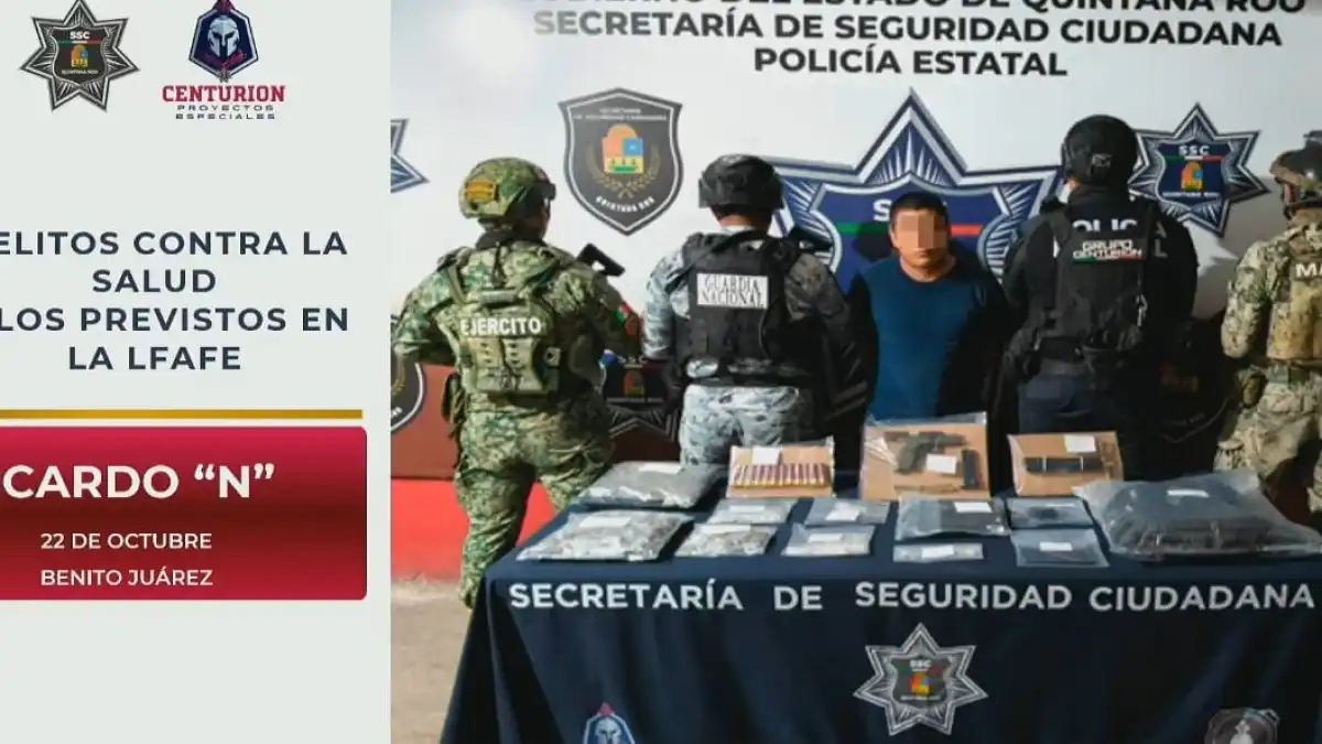 A group of armed personnel in military and police gear stand behind a table displaying seized items, with a masked individual in the center and official logos on a backdrop.