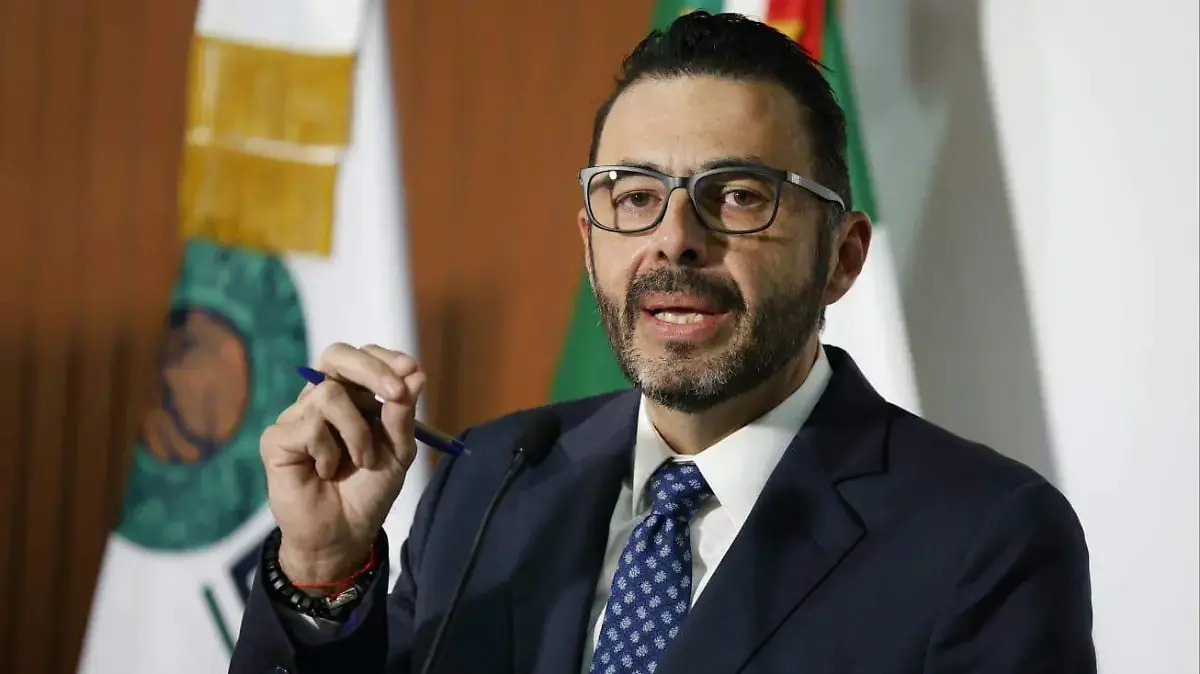 A man in formal attire speaks at a press conference, holding a pen and gesturing with his hand. Flags are visible in the background.