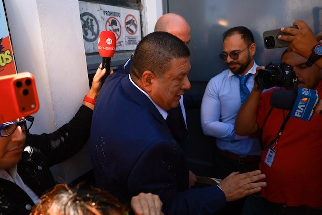 A man in a suit surrounded by journalists and cameras at a press event