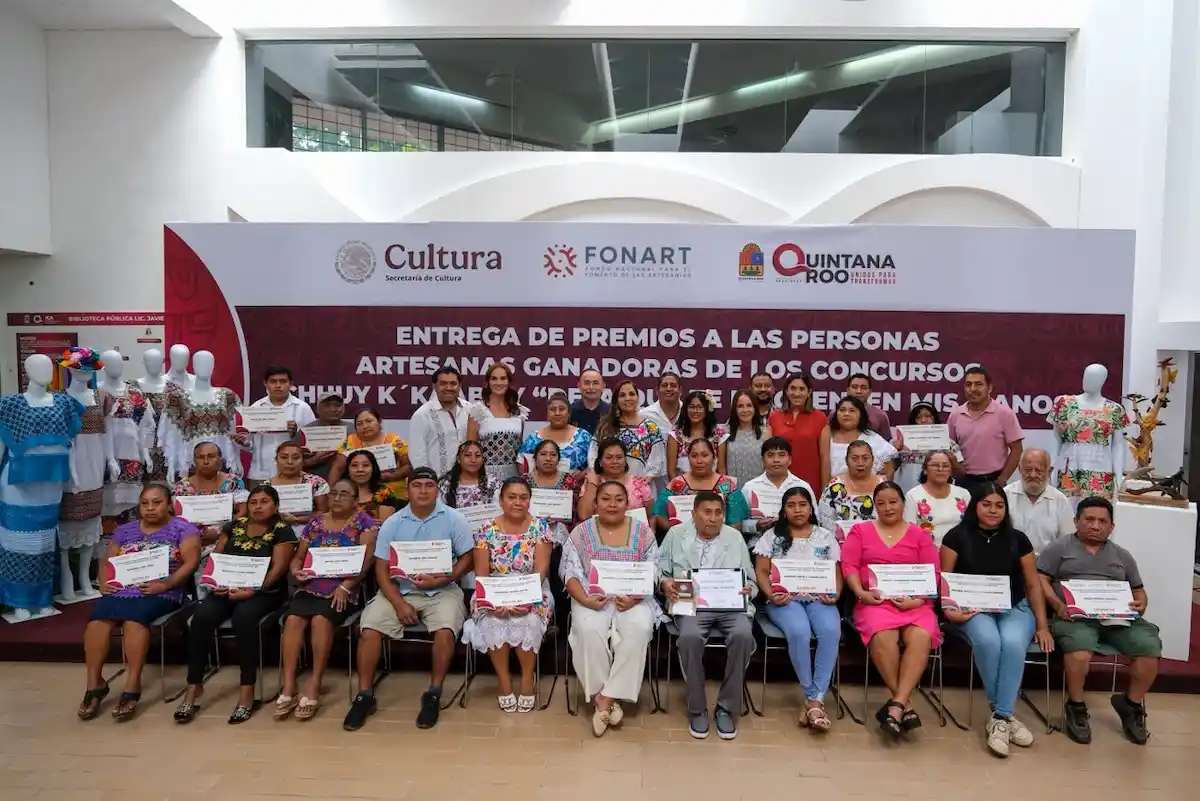 Group of artisans holding certificates at an award ceremony, standing in front of a banner and display of traditional clothing.$# CAPTION
