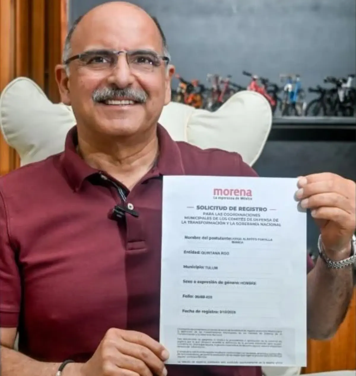 A man holding a registration application form for a political position, smiling at the camera in a home setting.$#$ CAPTION