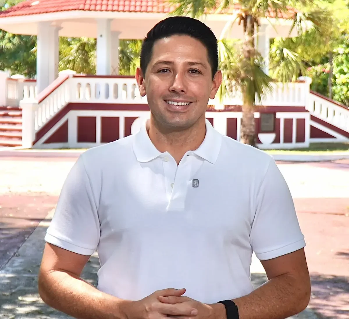 A smiling man in a white polo shirt stands outdoors in front of greenery and a decorative structure in the background.$# CAPTION