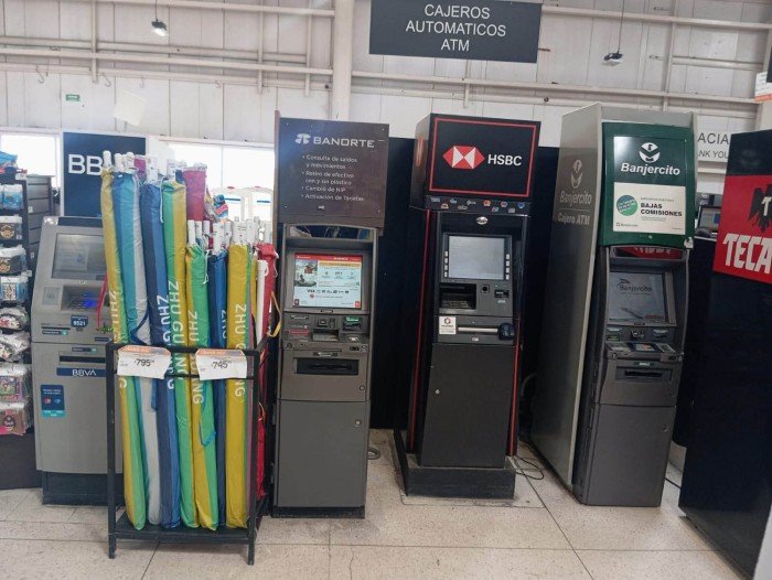 A row of four bank ATMs labeled BBVA Banorte HSBC and Banjercito stands beside a rack of folded umbrellas in a tiled indoor area under a sign reading Cajeros Automaticos ATM.