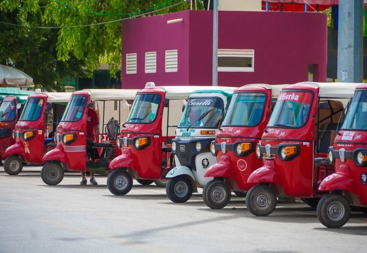 A line of red and white auto rickshaws parked on a paved surface in front of a magenta building with trees on the left.
