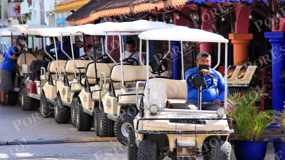 A line of white canopy golf carts is parked along a paved street beside colorful buildings, with a man in a blue shirt and face mask seated in the front cart.