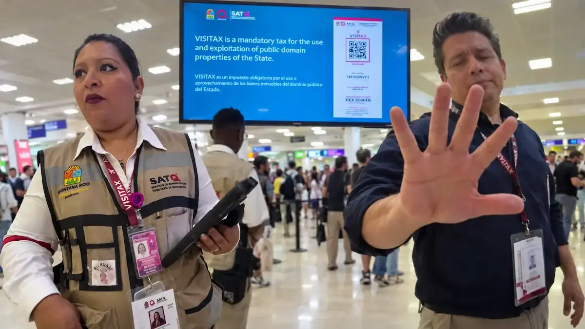 Two people wearing SATQ identification lanyards stand in an airport terminal in front of a large blue screen displaying VISITAX tax information and a QR code, with a woman holding a handheld device on the left and a man raising his hand on the right.