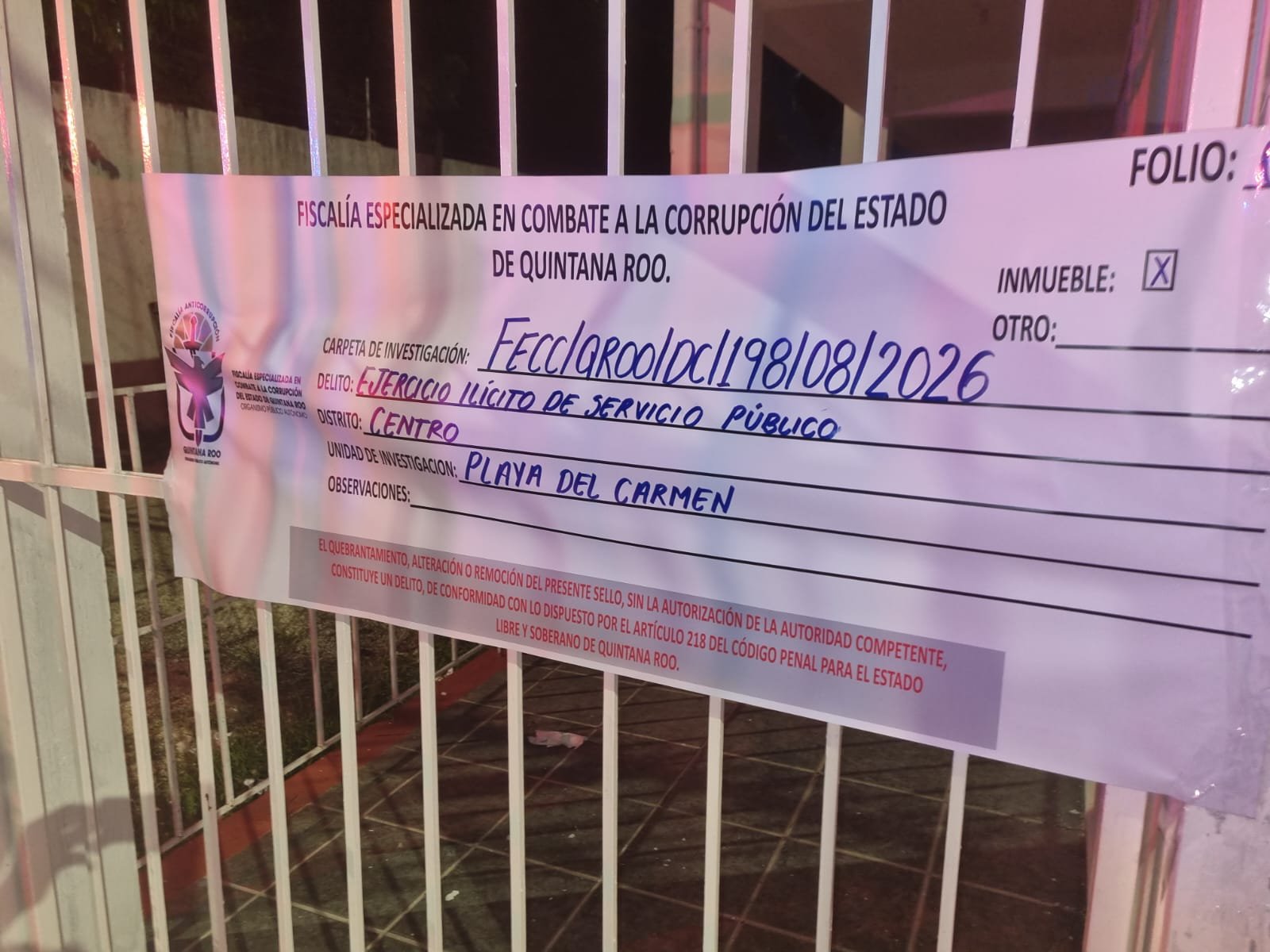 A printed official notice from the Quintana Roo anti-corruption prosecutor's office is attached to a white metal gate, with handwritten fields for investigation file, crime, district and investigating unit.