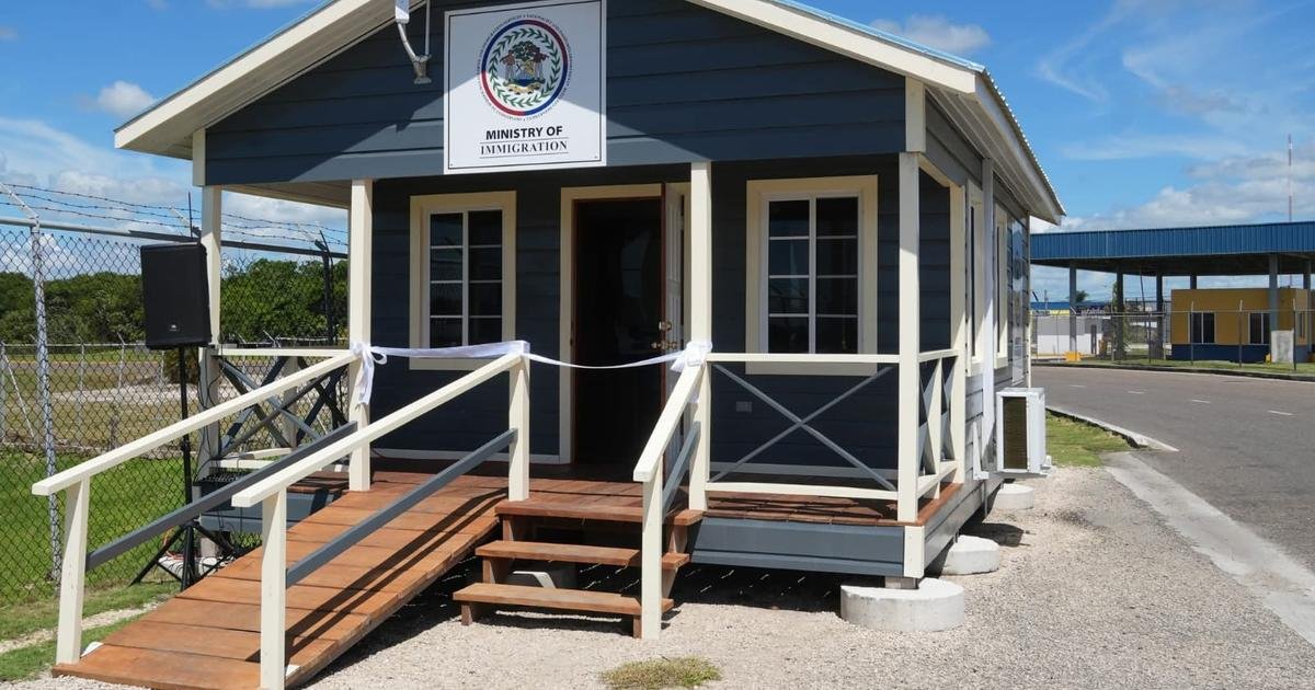 A small dark blue wooden building with white trim, a wooden ramp and steps, and a sign above the door reading Ministry of Immigration.