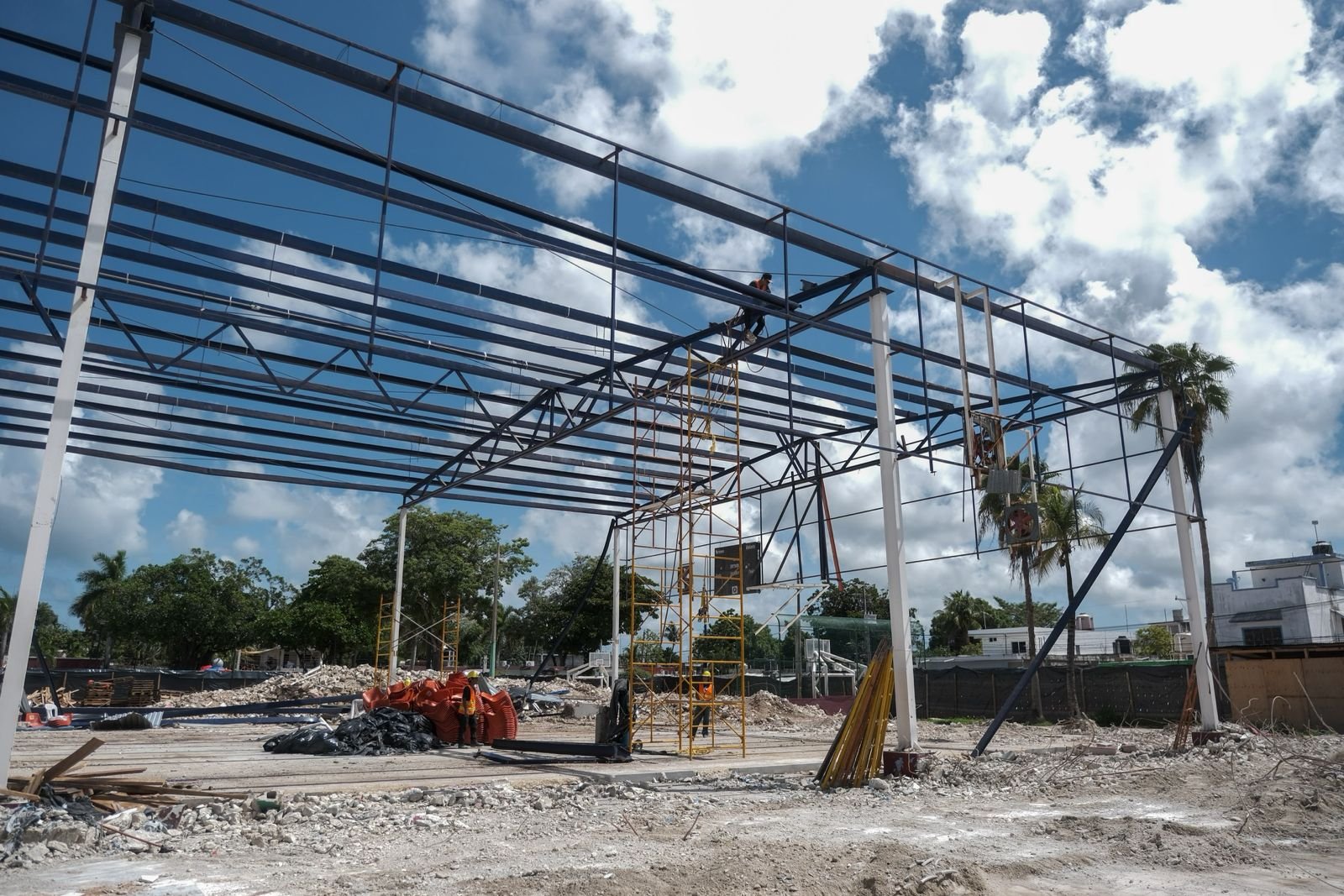 A large steel framework of a building under construction stands against a cloudy blue sky, with a worker on the upper beams, scaffolding, and construction materials on the ground below.