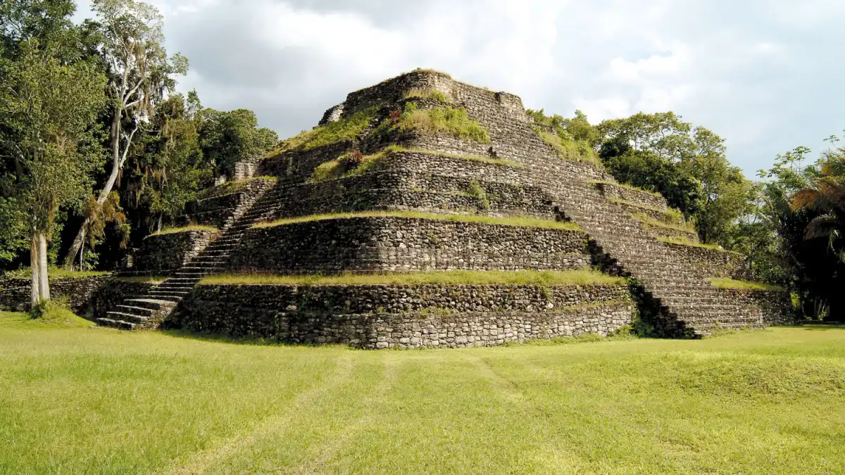 A large stepped stone pyramid covered in patches of grass and vegetation stands in a grassy clearing surrounded by trees under a cloudy sky.