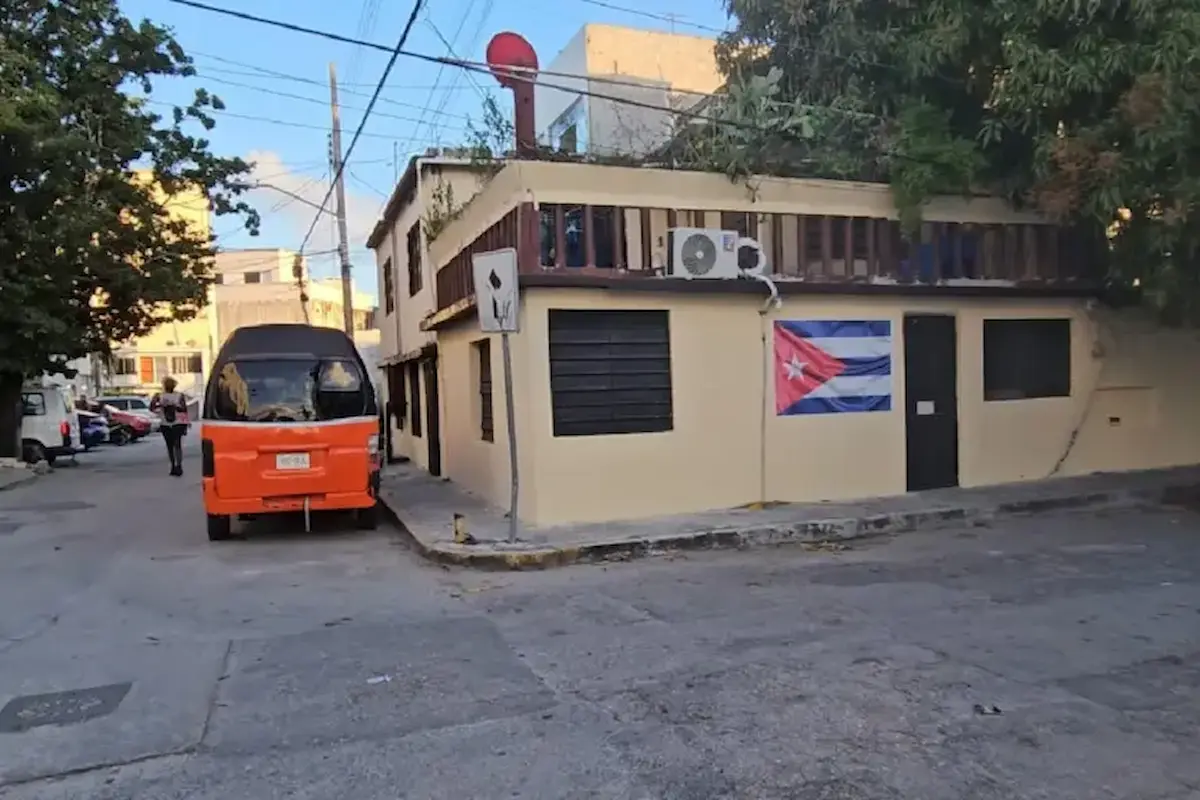 A street view featuring an orange vehicle parked beside a house with a Cuban flag on the wall, and a person walking in the background.$# CAPTION