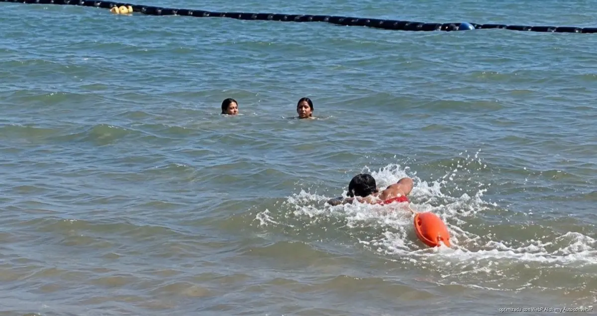 A person swims through choppy water pulling an orange rescue buoy while two other people watch from behind, with a dark floating line across the top of the frame.