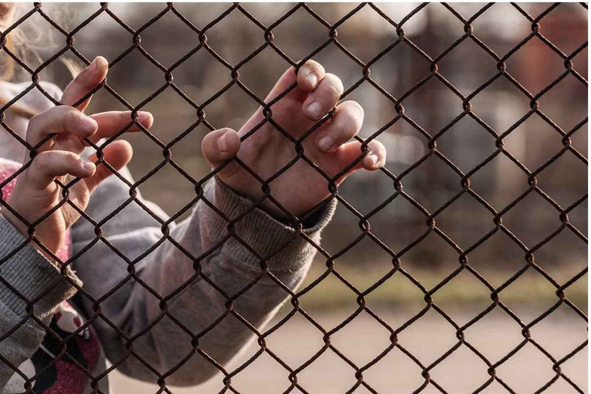 Close-up of two hands grasping a chain-link fence, with a blurred background of an open field.$# CAPTION