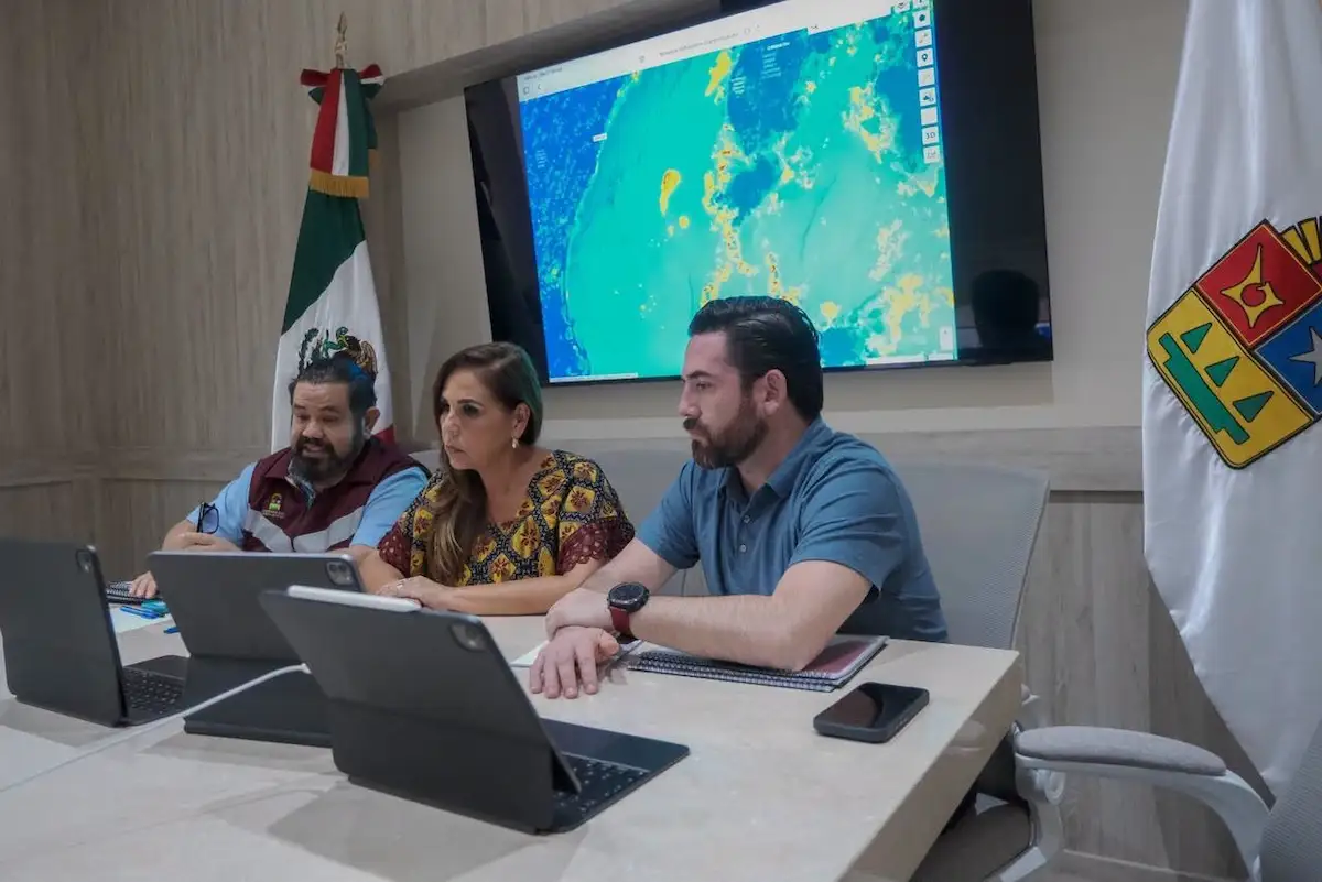 Three people sit at a table with laptops in front of a large screen displaying a colorful weather map, with a Mexican flag on the left and a white flag with a crest on the right.