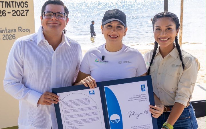 Three smiling people stand on a sandy beach holding a large open certificate folder, with a person wading in the water behind them.