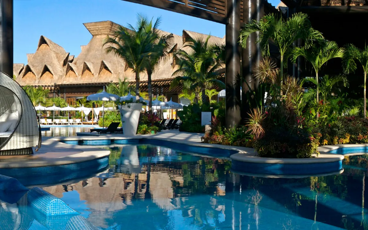 A serene pool area surrounded by lush tropical greenery and modern architecture, including thatched roofs and lounge chairs under umbrellas