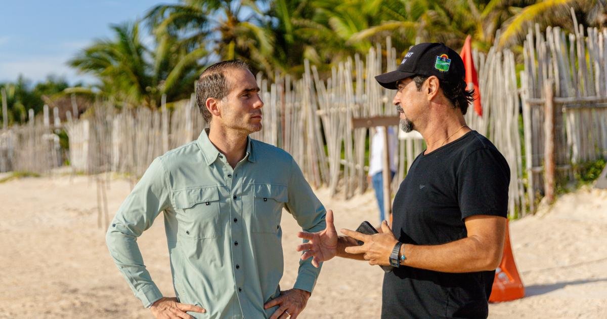 Two men stand facing each other on a sandy beach in front of a wooden stick fence and palm trees, one wearing a light green button shirt and the other a black t shirt and black cap.