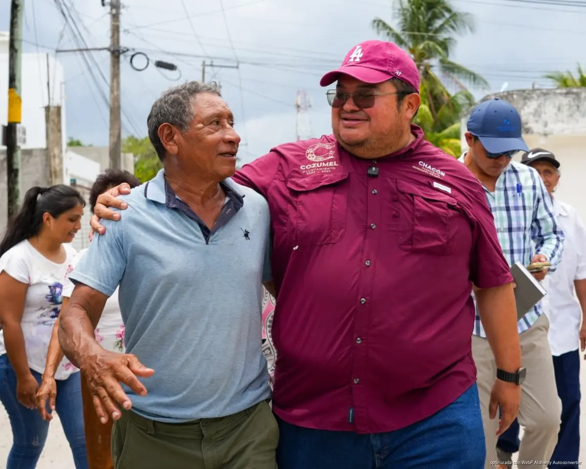 A man in a maroon shirt and cap with his arm around another man in a light blue polo shirt as they walk on a street with other people behind them.