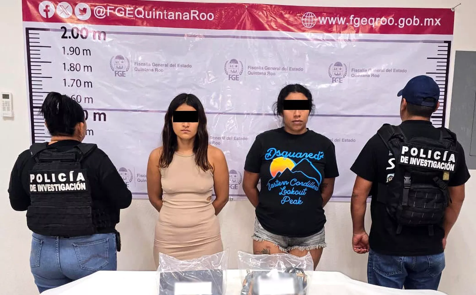 Two women with black bars over their eyes stand in front of a height chart banner between two officers wearing Policia de Investigacion vests, with plastic-wrapped items on a table in front.