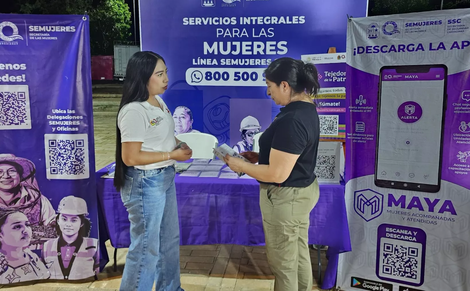 Two women stand facing each other in front of purple Semujeres banners and a table with informational materials, one holding a pamphlet.