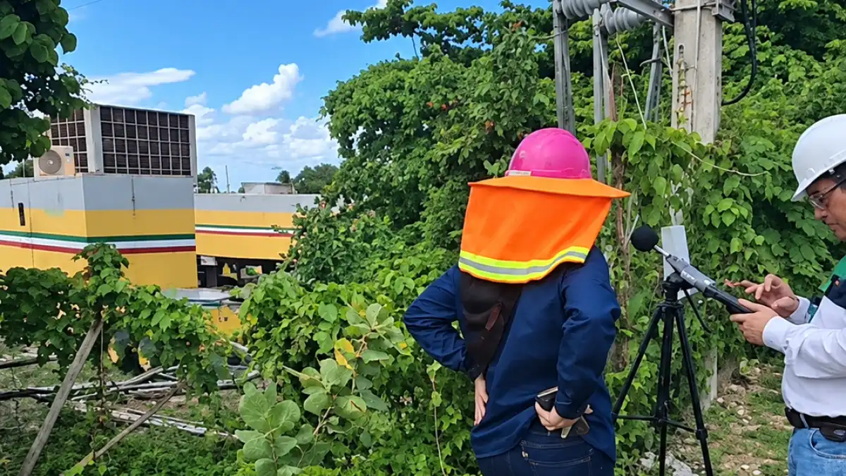 Two people in hard hats stand outdoors beside a tripod mounted device, with a yellow and grey building and dense green foliage behind them.