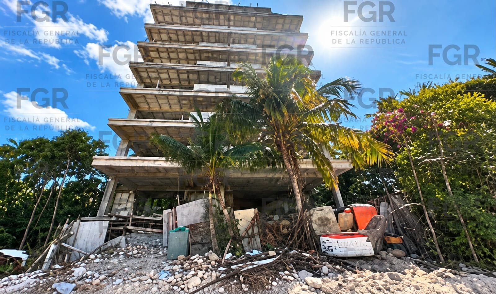 An unfinished multi-story concrete building with exposed floor slabs stands behind palm trees and a pile of rubble and debris on a rocky ground under a blue sky, with FGR and Fiscalia General de la Republica watermarks across the image.