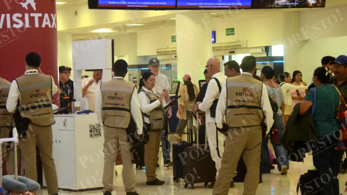 Several workers wearing tan Visitax vests stand near a white counter and travelers with luggage in an airport terminal, with a red Visitax sign on the left and flight information screens above.