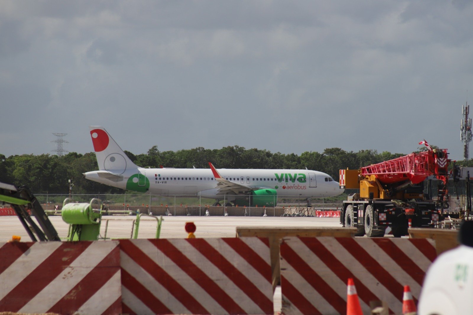 A white Viva Aerobus Airbus passenger jet with green and red tail markings sits on an airport tarmac behind red and white striped barriers, with a red crane truck to the right and trees under a cloudy sky in the background.