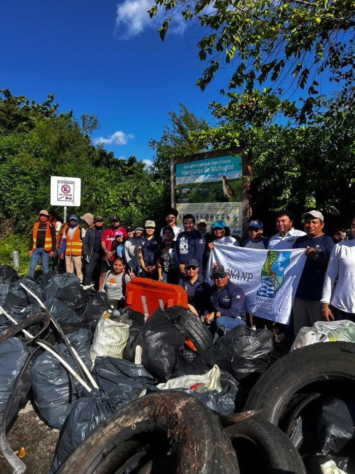A group of volunteers stands behind large piles of black garbage bags and discarded tires in front of a green Manglares de Nichupte nature reserve sign.