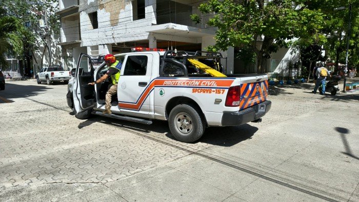 A white pickup truck marked Proteccion Civil with orange stripes is parked on a paved street with its driver door open and a person in a red helmet and yellow vest stepping out.