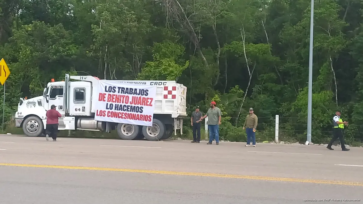 A white dump truck with a large banner reading LOS TRABAJOS DE BENITO JUAREZ LO HACEMOS LOS CONCESIONARIOS is parked on the shoulder of a paved road beside dense green trees, with several men standing nearby and one person in a yellow vest walking on the road.
