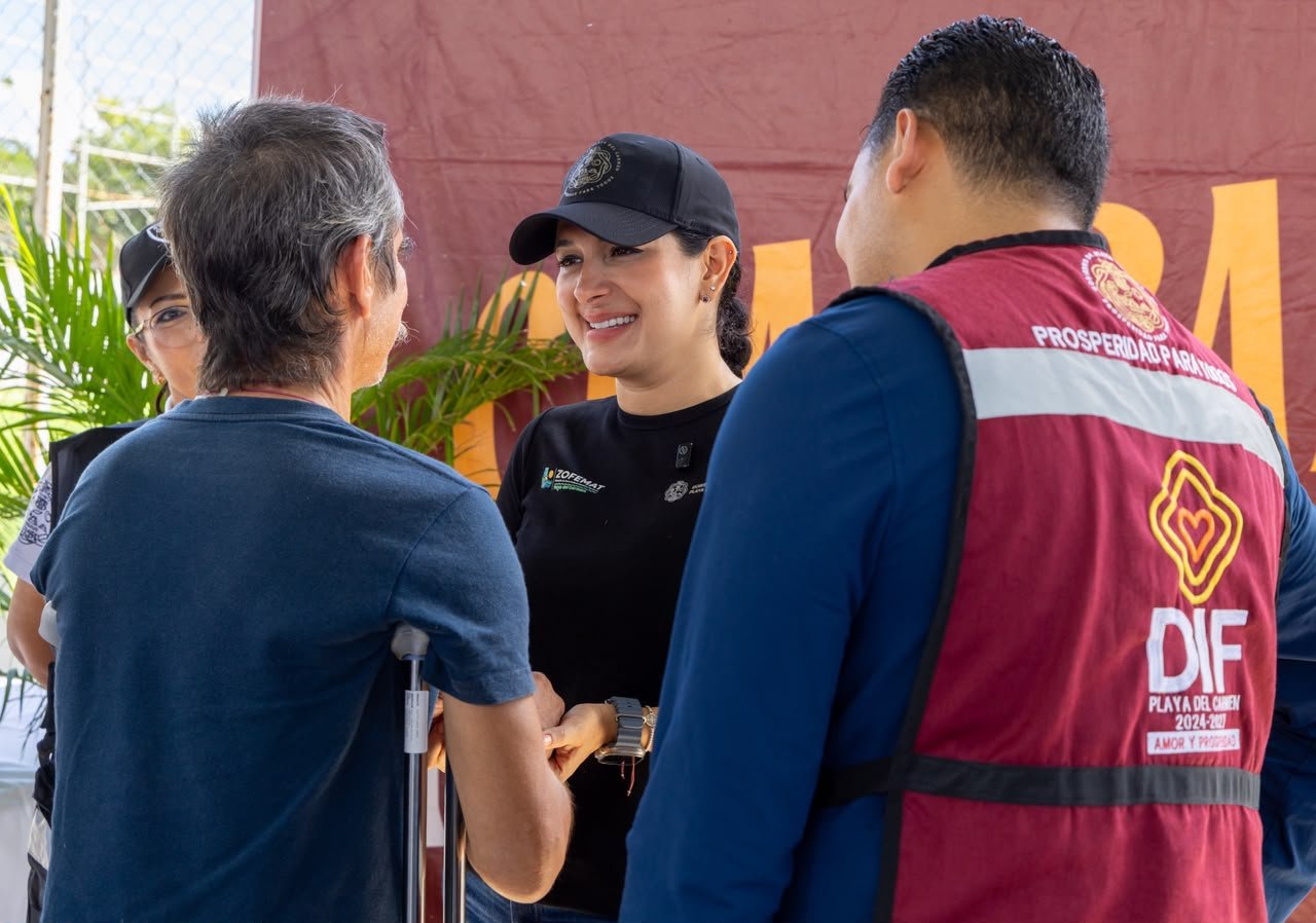 A smiling woman wearing a black cap and black shirt stands facing a man in a blue shirt who holds a crutch, while a person in a red DIF vest stands to the right and another person is partially visible on the left.
