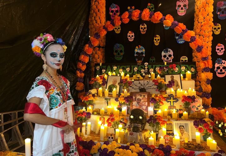 A woman with sugar skull face paint and a flower headdress stands beside a large Day of the Dead altar decorated with marigolds, candles, paper skulls and framed photographs.