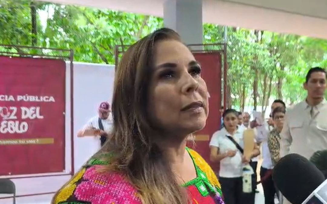 A woman with long brown hair wearing a brightly embroidered dress stands outdoors in front of a red banner and several people, with a microphone visible in the lower right corner.