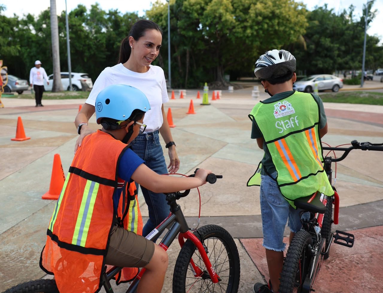 A smiling woman in a white t-shirt stands between two children wearing helmets and safety vests who are seated on bicycles on a paved area marked with orange traffic cones.