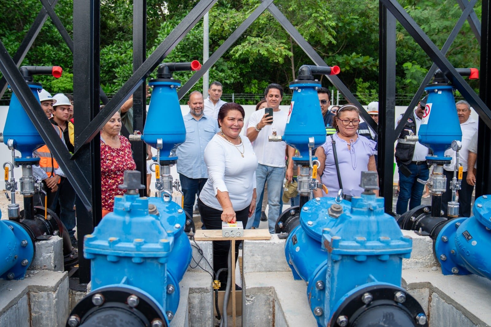 A woman in a white top presses a control box mounted on a wooden plank between large blue industrial valves as a group of people watches behind her.