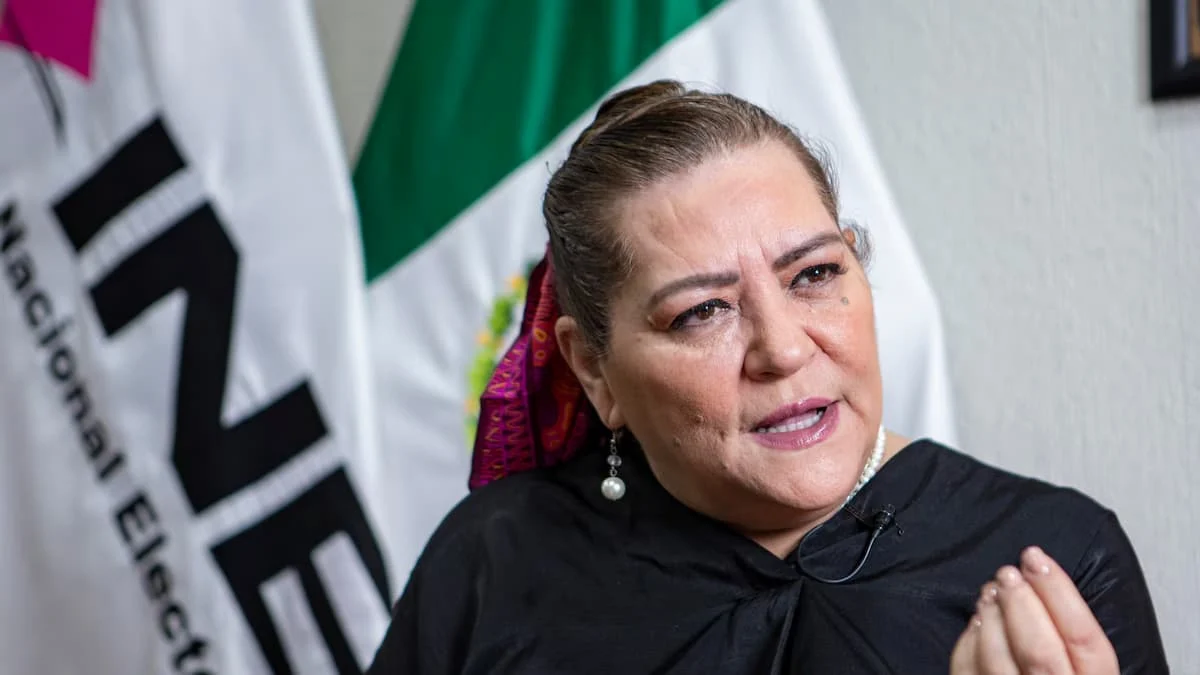 A woman in a black top with a pearl earring and a small lapel microphone speaks in front of a white INE banner and a Mexican flag.