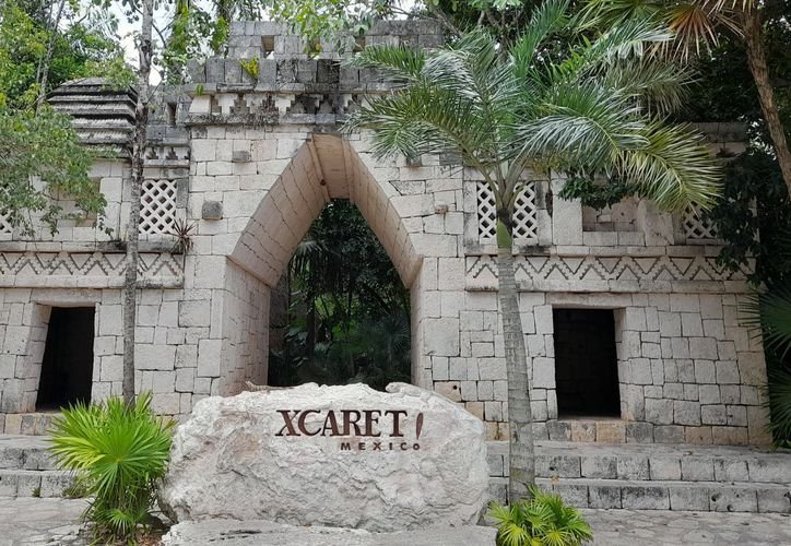 A large stone archway with a pointed top stands behind a rock sign reading XCARET MEXICO, surrounded by palm trees and tropical greenery.